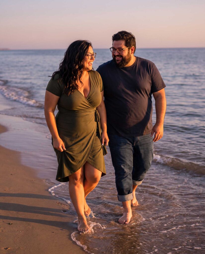 Anna und Karl laufen barfuß am Strand entlang und schauen sich lächelnd an.
